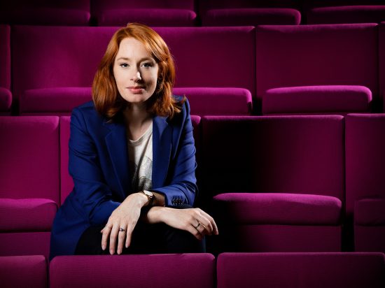 Image of Hannah Fry sitting on a row of purple auditorium seats, arms folded over her knees. 
