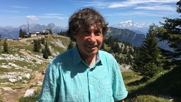 Paul, wearing an aqua shirt, smiles toward the camera. Rocky land and a bright blue sky are visible in the background.
