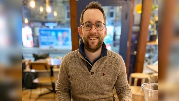 Nathan, wearing a light brown sweater, smiles toward the camera. The interior of a cafe is visible in the background.