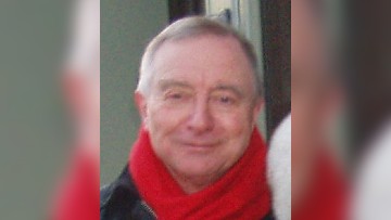 A head and shoulders shot of Russel, wearing a red scarf and black jacket, smiling toward the camera. The background is blurred.