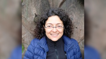 Sara smiles toward the camera. She has curly black hair and is wearing a black fleece and a dark blue coat. She is standing against a wooden fence.
