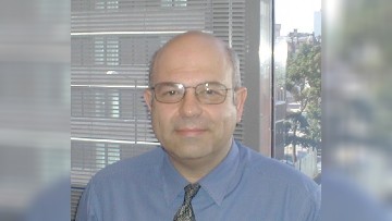 Aki, wearing a blue shirt and patterned bronze tie, smiles towards the camera. Behind him, closed blinds are visible to the left, and trees and buildings to the right.