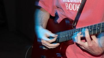 A man in a faded red tshirt plays a burgundy electric guitar. Only his mid-section and hands / arms are visible.
