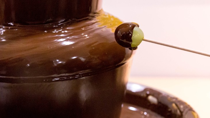 A chocolate fountain. A skewer holding a chocolate-covered grape is on the right hand side of the image.