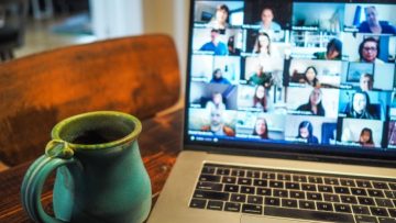A laptop with a video conference in progress on the screen. A green coffee cup is on the desk to the left of it.