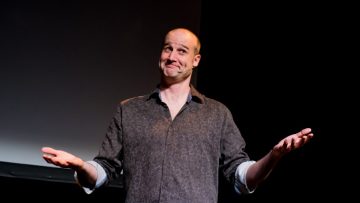 Matt stands in front of a black background wearing a black shirt. He is smiling with his mouth closed and holds his hands up as if shrugging