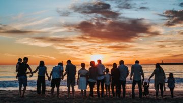 A group of adults and children standing in a line on a beach with their backs to the camera. They are watching a sunrise.