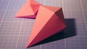 Two pieces of origami made with red-orange paper. They are laying on a gridded blue cutting mat.