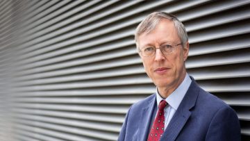 Nicholas-Higham looking towards the camera. He is wearing a navy suit, blue shirt and red tie, and is standing against a corrugated metal wall.