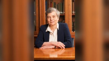 Caroline Series sits with her hands crossed on a desk in front of her. She is wearing a white blouse and navy blue jacket, and smiling toward the camera. She is sitting in front of a wooden bookshelf.