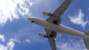 A passenger airplane in flight, viewed from below. The sky is blue with white, whispy clouds.