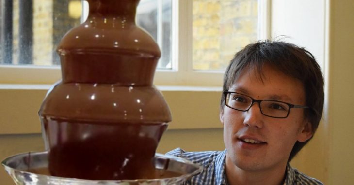 Adam Townsend is crouched beside a chocolate fountain, looking at the chocolate cascading down.