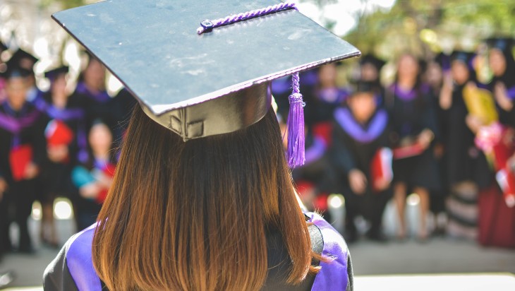 The head and shoulders of a long-haired female graduate. A group of graduates are visible in front of her, out of focus