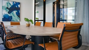 A white-walled conference room with one glass wall, There is an abstract painting in shades of blue on the white wall. There is an oval table with a pot plant on, surrounded by tan leather chairs.