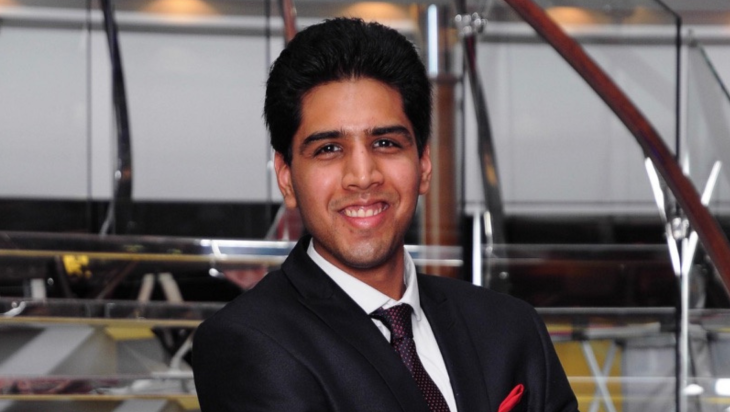 Rehan Shah, dressed in a black suit and tie, faces the camera, smiling
