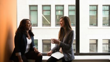 Two females are smiling at eachother in conversation, sitting by a window in a multi-storey building.